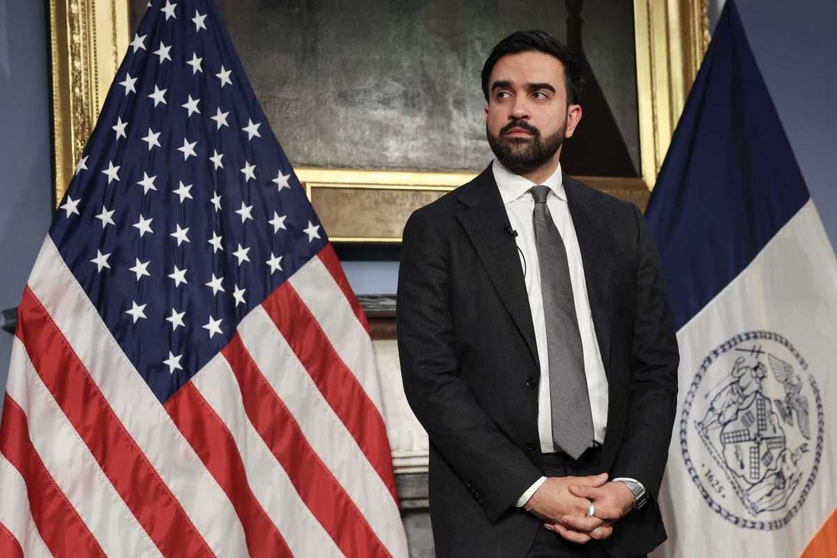 Zohran Mamdani, a bearded brown man with short black hair, stands in front of two flags, looking to his right.