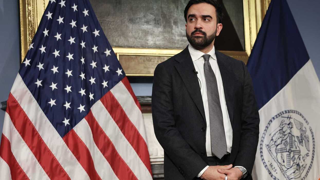 Zohran Mamdani, a bearded brown man with short black hair, stands in front of two flags, looking to his right.