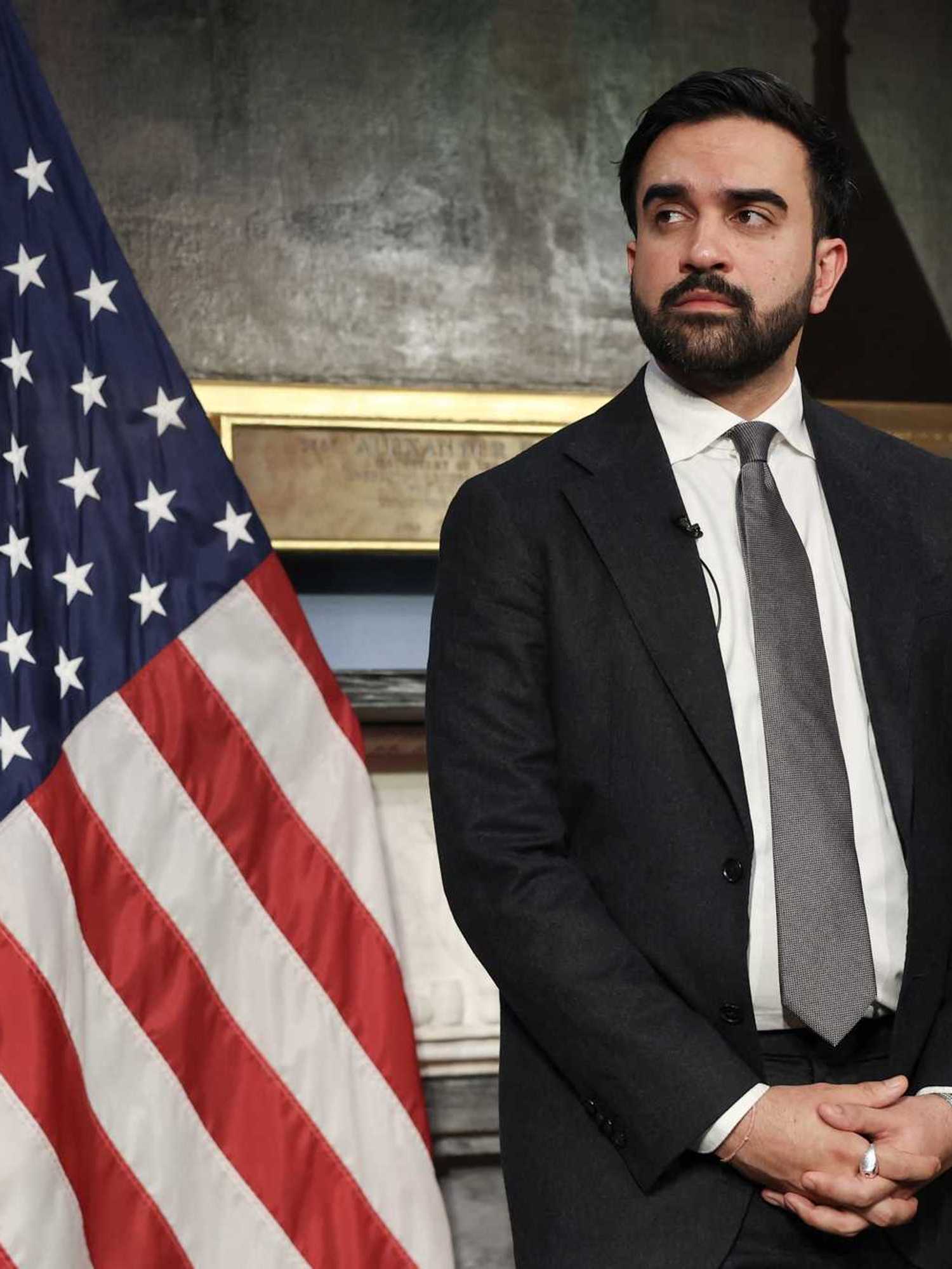 Zohran Mamdani, a bearded brown man with short black hair, stands in front of two flags, looking to his right.
